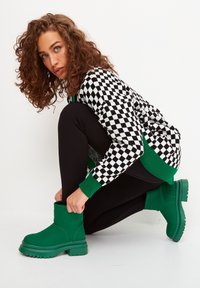 Zöld bokacipők masszív gumitalppal, sima textúrával és egyszerű designnal. Fekete leggingssel és fekete-fehér kockás pulóverrel viselve.
