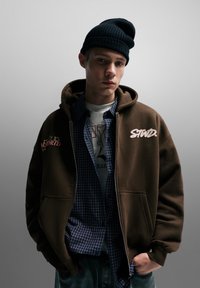 Brun hættetrøje med lynlås og broderede stjerner og logo, lagdelt over en blåternet skjorte. Afsluttet med en sort beanie-hue. Casual streetwear-stil.