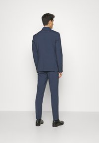 Marineblauer Anzug mit einreihigem Jackett und maßgeschneiderten Hosen. Glatte Textur, abgerundeter Kragen und geknöpfte Manschetten. Schwarze Schuhe.