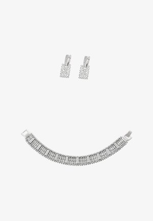 Ezüst karkötő bonyolult, texturált láncszemekkel és apró gömb alakú díszítésekkel. Párban viselve, hozzáillő ezüst fülbevalókkal, melyeken dekoratív minták találhatók.