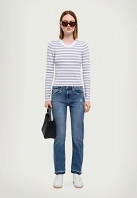 Maglietta a maniche lunghe a righe grigio chiaro e bianco, jeans blu, sneakers bianche e borsa nera. La modella indossa occhiali da sole scuri.