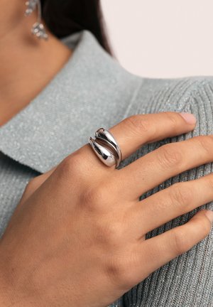 Anillo de tono plateado con un diseño en espiral. Superficies lisas y reflectantes. Llevado en una mano contra un suéter texturizado de color azul claro.