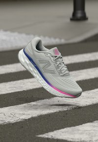 Chaussure de sport gris clair avec tige en mesh, accents argentés et une semelle intermédiaire dégradée colorée passant du violet au rose. Semelle texturée pour une meilleure adhérence.
