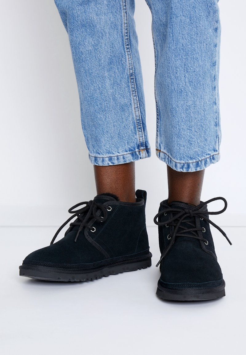 Bottines en suède noir avec bout rond, semelles en caoutchouc plates et détail à lacets. Portées avec un jean bleu clair retroussé.