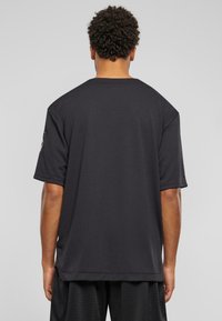 Tricou sport negru, cu mânecă scurtă, cu o croială lejeră, având un guler rotund și o textură subtilă. Include un accent cu logo lângă umăr.