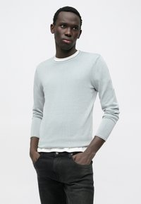 Ανοιχτό γκρι πλεκτό πουλόβερ με μακριά μανίκια, στρογγυλή λαιμόκοψη και διακριτική υφή, σε συνδυασμό με σκουρόχρωμο στενό τζιν. Μινιμαλιστικός σχεδιασμός χωρίς σχέδια.