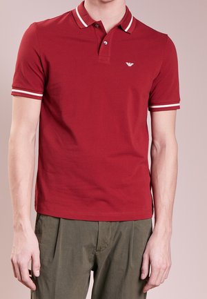 Homme portant un polo rouge à manches courtes avec un liseré blanc au col et aux manches, associé à un pantalon vert olive.
