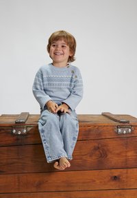 Helles blaues Sweatshirt mit dekorativen Mustern und drei braunen Knöpfen am Ausschnitt, kombiniert mit hellblauen Jeans. Kind sitzt barfuß.