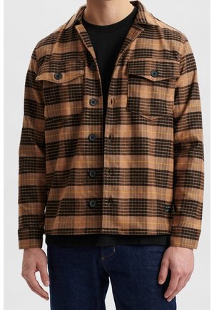 Chaqueta de cuadros marrón y negra con acentos amarillos, que cuenta con dos bolsillos en el pecho, botones y una textura suave. Llevada sobre una camisa negra.