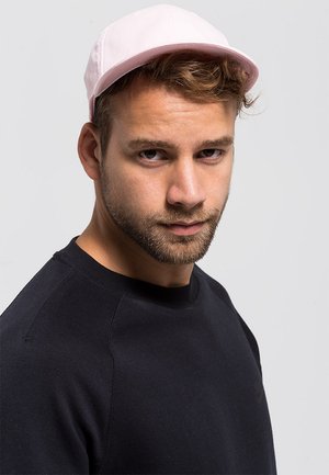 Șapcă roz din țesătură cu un cranium rotund și vizieră rigidă, purtată împreună cu un hanorac negru cu un logo subtil.