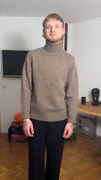 Brown, chunky knit sweater with a tall turtleneck, long sleeves, and ribbed hem, paired with black, wide-leg pleated trousers.