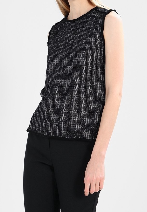 Černý bezrukávový top s texturovaným kostkovaným vzorem, s kulatým výstřihem a třepenými okraji. V kombinaci s černými kalhotami, které ukazují přiléhavý siluetu.