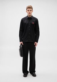 Μαύρο πουκάμισο με κουμπιά και μικρή κόκκινη ετικέτα, συνδυασμένο με φαρδιά μαύρα παντελόνια. Το μοντέλο κρατάει μια μαύρη τσάντα χαρτοφύλακα. Απλός, μοντέρνος σχεδιασμός.