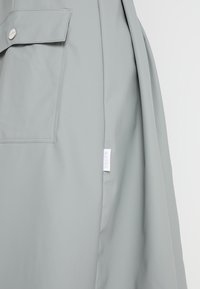 Jasnoszary płaszcz o gładkiej fakturze, z kieszenią na piersi i małą metką marki na bocznym szwie. Minimalistyczny design i czyste linie.