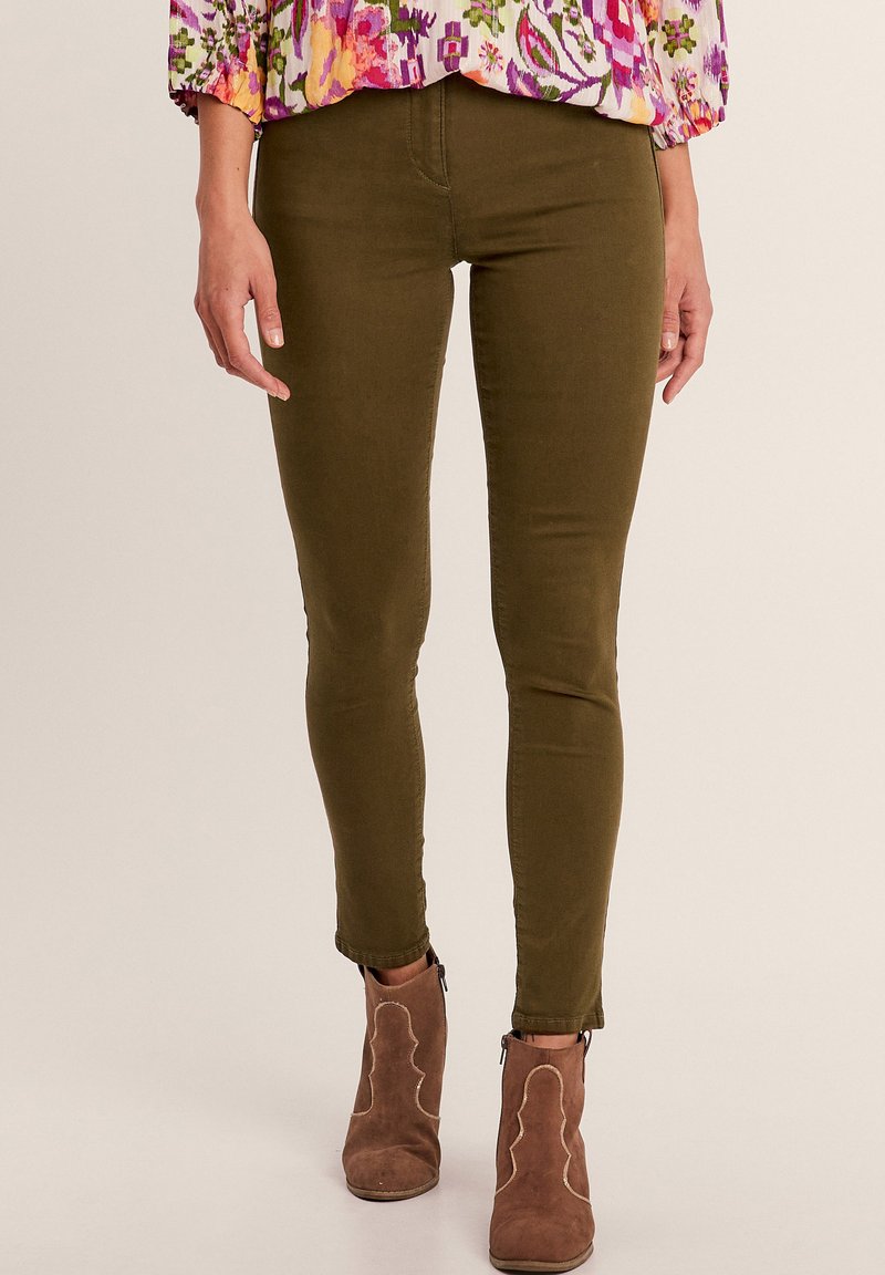 Jeans skinny verde oliva con una texture liscia e una vestibilità affusolata, abbinati a stivaletti marrone chiaro con cuciture decorative.