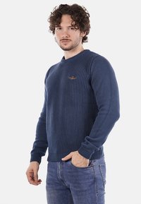 Maglione lavorato a maglia blu navy con un motivo testurizzato, polsini e orlo a coste, e un piccolo logo sul petto, abbinato a jeans in denim blu.