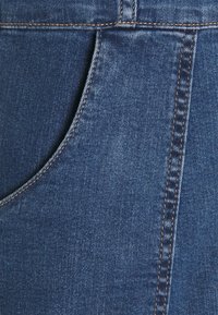 Närbild av blå denimtyg som visar framfickan och den vertikala sömmen med orange sömmar.