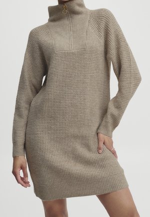 Beige wafelgebreide sweaterjurk met lange mouwen en een hoge kraag met rits, gedragen door een persoon met één hand op de heup.