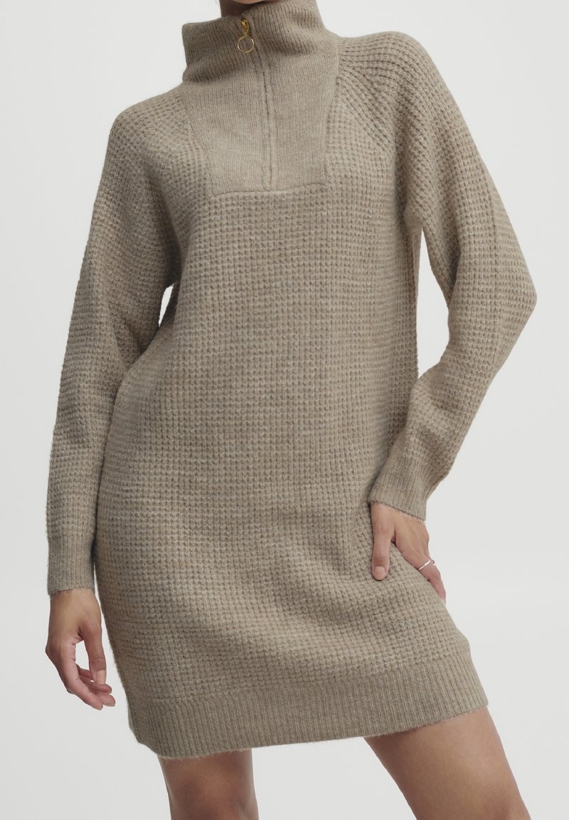 Robe-pull beige en maille gaufrée avec manches longues et col montant zippé, portée par une personne avec une main sur la hanche.