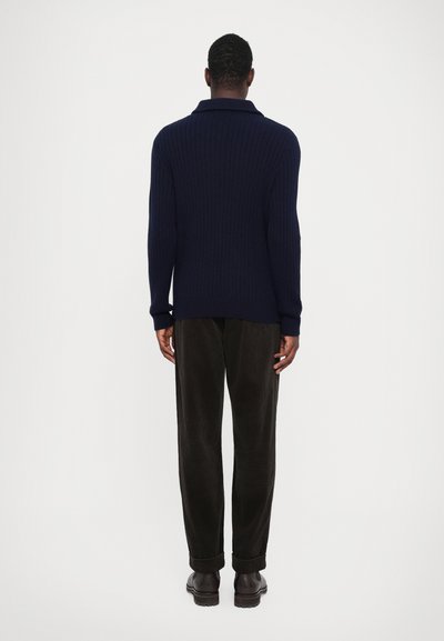 Suéter de punto acanalado azul marino con un cuello doblado, combinado con pantalones de pana oscuros y zapatos marrones resistentes, visto desde atrás.
