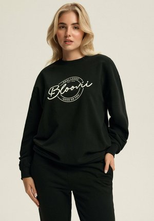 Czarny sweatshirt wykonany z miękkiego materiału, z białym okrągłym logo z tekstem na przodzie. Długie rękawy i luźny krój.