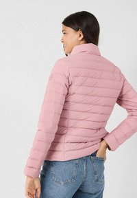 Chaqueta acolchada de color rosa claro con detalles de costura vertical, cuello alto y mangas ajustadas, combinada con jeans de mezclilla. Textura suave.