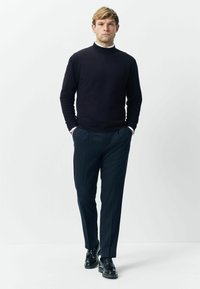 Navy turtleneck sweater paired with dark textured trousers. Model stands with hands in pockets, wearing black shoes and white shirt cuffs visible.