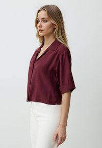 Burgunderfarbene Bluse mit kurzen Ärmeln und Kragen, mit durchgehender Knopfleiste, glatter Textur und verkürztem Design, kombiniert mit weißen Hosen.