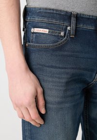 Tmavomodré džínsy s úzkym strihom, s viditeľnou nálepkou na zadnej vrecku, meďou šitím a minimálnym detailom.