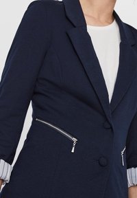 Marinblå kavaj med spetsig krage, två fickor med dragkedja på sidorna och knappstängning. Inuti finns en ljusfärgad foder och randiga ärm detaljer.