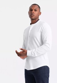 Camisa blanca de manga larga con cuello redondeado y cierre de botones en la parte frontal. Textura suave, corte slim, combinada con unos jeans azul oscuro.