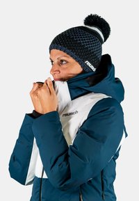 Navy blue jacket with white accents and zippers, paired with a knitted dark blue beanie featuring a white stripe and pompom on top.