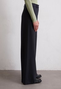 Schwarze, weit geschnittene Hosen mit glatter Textur und taillierter Passform. Verfügt über einen flachen Bund, Gürtelschlaufen und Seitentaschen.
