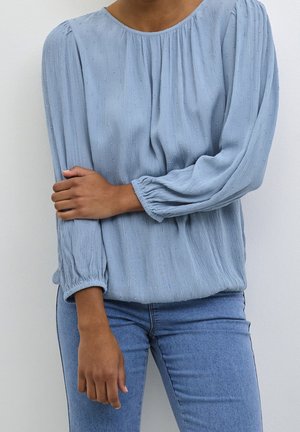 Person wearing a light blue textured long-sleeve blouse with elastic cuffs and blue jeans, standing against a plain white background.