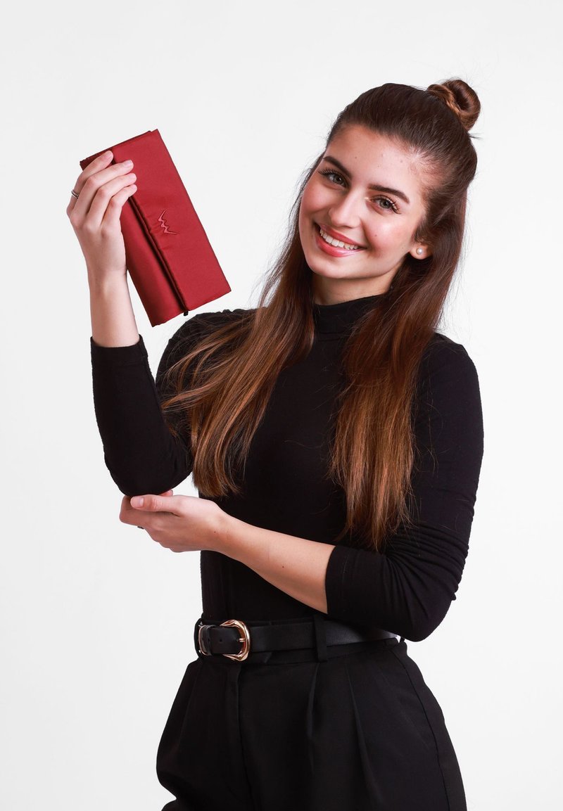 Red wallet with an embossed zigzag pattern, made of smooth leather, held in a hand against a plain white background. Black long-sleeve shirt.