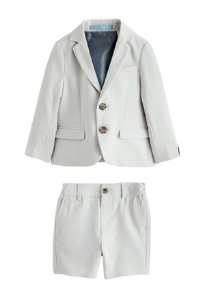 Jeune garçon debout et souriant, portant un blazer gris clair, un short assorti, une chemise blanche et des mocassins beiges sur un fond uni.