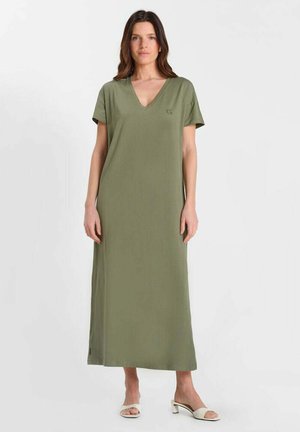 Mujer de pie vistiendo un vestido maxi de manga corta y escote en V de color verde oliva, y sandalias blancas de tacón abierto, sobre un fondo liso.