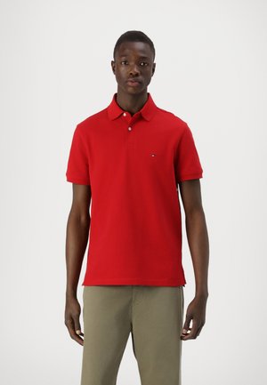 Rød polo skjorte med klassisk krage, kort ermer, og en liten merke logo på brystet. Kombinert med lyse kaki bukser.