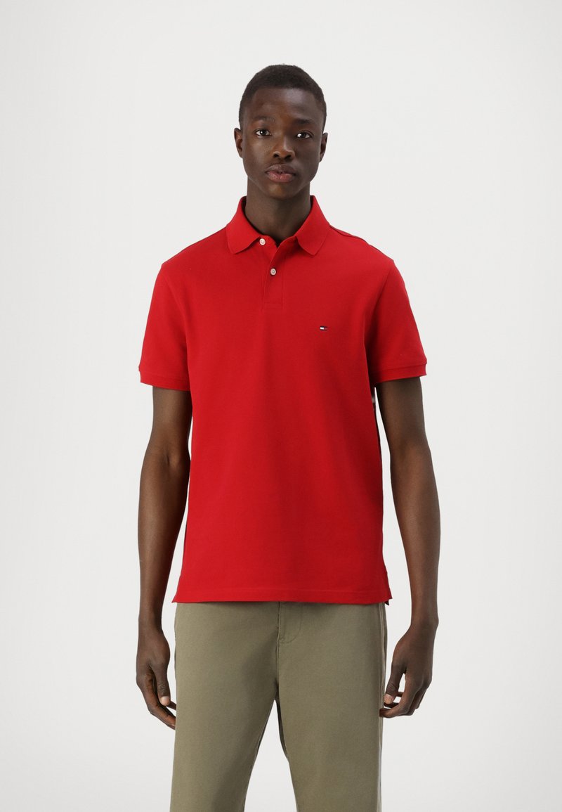 Camisa polo roja con cuello clásico, mangas cortas y un pequeño logo de la marca en el pecho. Combinada con pantalones de caqui claros.