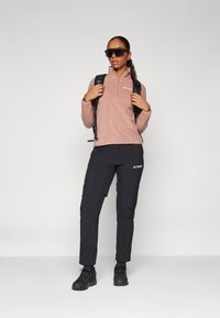 Rosa Fleece-Halzip-Pullover, schwarze elastische Hose mit Taschen, robuste schwarze Schuhe und große schwarze Sonnenbrille, dazu ein schwarzer Rucksack.