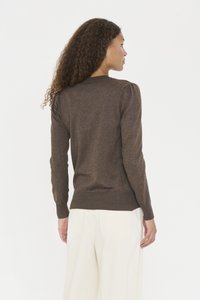 Femme aux cheveux bouclés portant un pull marron ajusté et un pantalon blanc ample, de dos contre un fond uni.