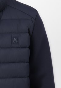 Mornarsko prsluk s punjenjem, glatke površine, s kviltanim dizajnom, visokim ovratnikom i malom logotipom na prednjoj strani.