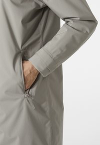 Světle šedá vodotěsná bunda s hladkým povrchem, postranní kapsa na zip a nastavitelné manžety. Vyznačuje se volným střihem.