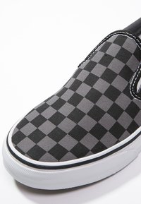Baskets slip-on à carreaux noirs et gris avec semelle en caoutchouc blanche et bordure noire, présentées en gros plan sur fond blanc.