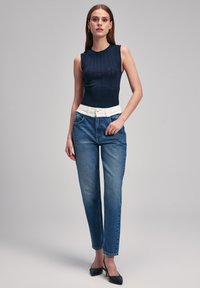 Top sin mangas de color navy con diseño de punto texturizado, combinado con jeans de mezclilla azul claro con cintura blanca y bolsillos laterales. Tacones negros.