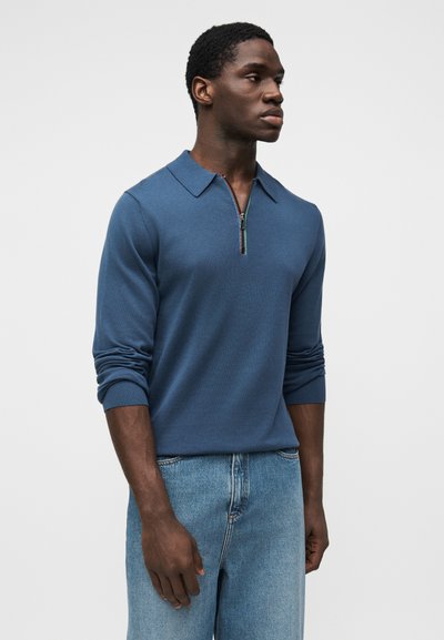 Νεαρός άνδρας που φοράει μπλε πουλόβερ με φερμουάρ και μακριά μανίκια και ανοιχτόχρωμο τζιν, στέκεται με τα χέρια χαλαρά σε λευκό φόντο.