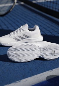 Pair of white Adidas tennis shoes on blue court surface, with one shoe showing detailed tread pattern in foreground.