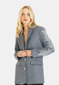 Blazer grigio con una vestibilità su misura, caratterizzato da un intricato ricamo argentato sulla manica sinistra, due tasche frontali e una chiusura con bottone a un solo petto.