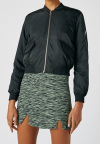 Veste bomber noire zippée portée avec une mini-jupe à rayures tigre verte et noire avec deux fentes sur le devant, montrée sur une personne des épaules aux cuisses.