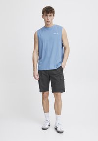 Cămașă fără mâneci, de culoare albastru deschis, cu logo, asortată cu pantaloni scurți negri tip cargo. Modelul poartă șosete albe și adidași gri și negri.